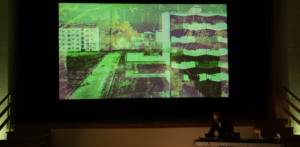 Images shows a scene from the Sz. Berlin performance Disonantna sinteza, June 2023. A digitally altered cityscape is visible on the screen, with the performer in front of it.